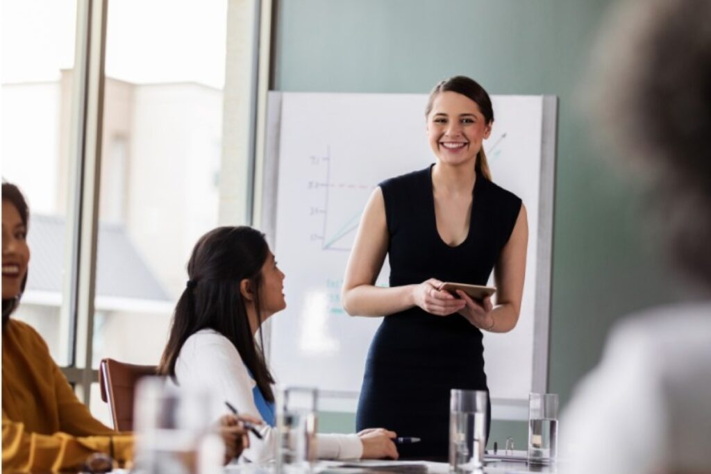 a young woman with dark brown hair is giving a presentation in front of a group of people. She appears confident and in contol.